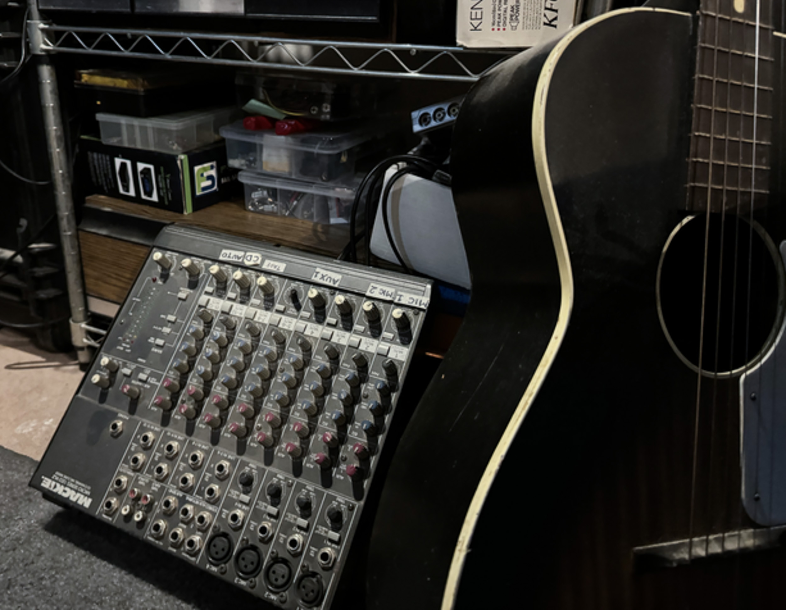 An audio mixing board and guitar in front of shelves.