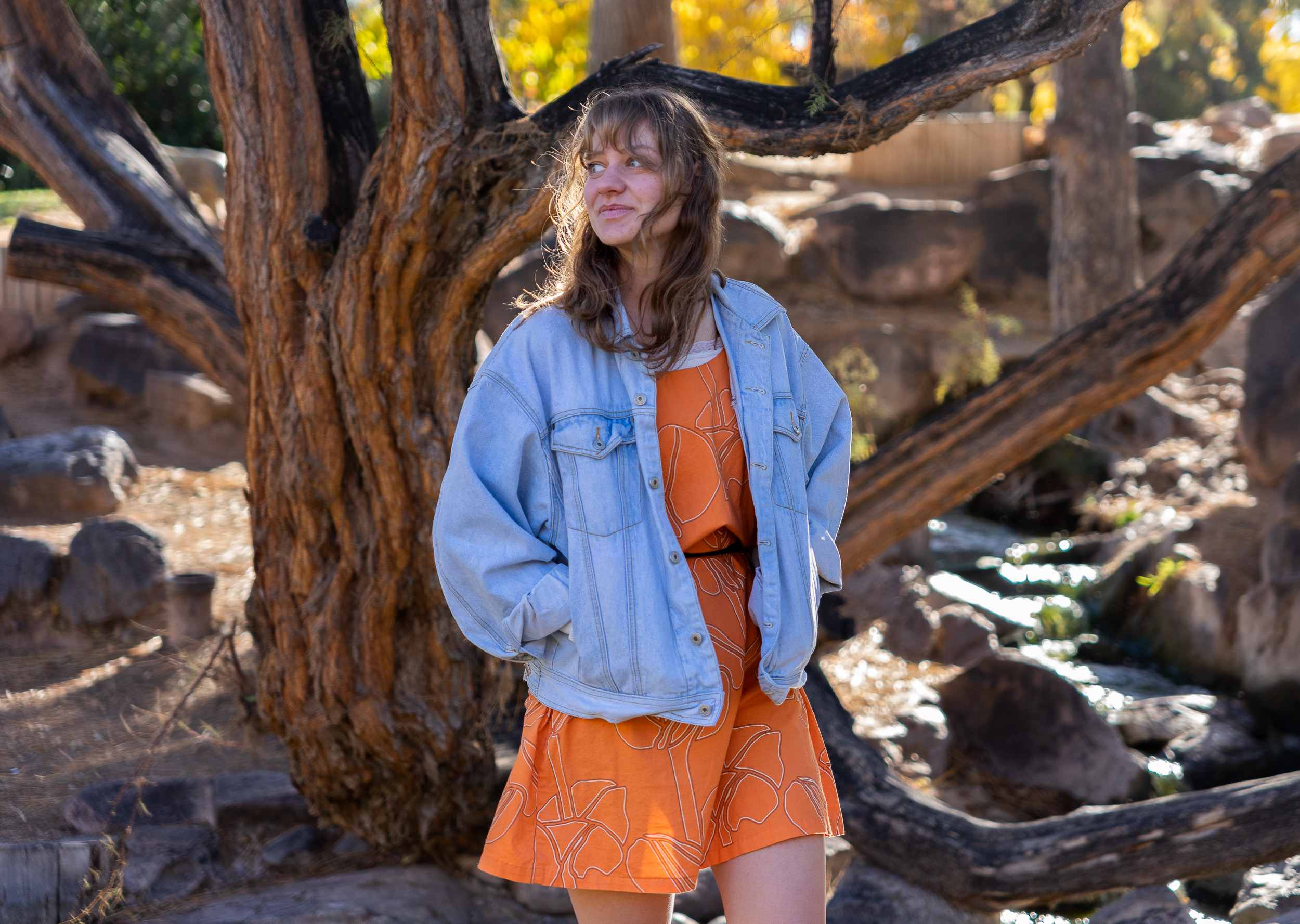 A woman wearing an orange dress and blue jacket stands beneath a tree.